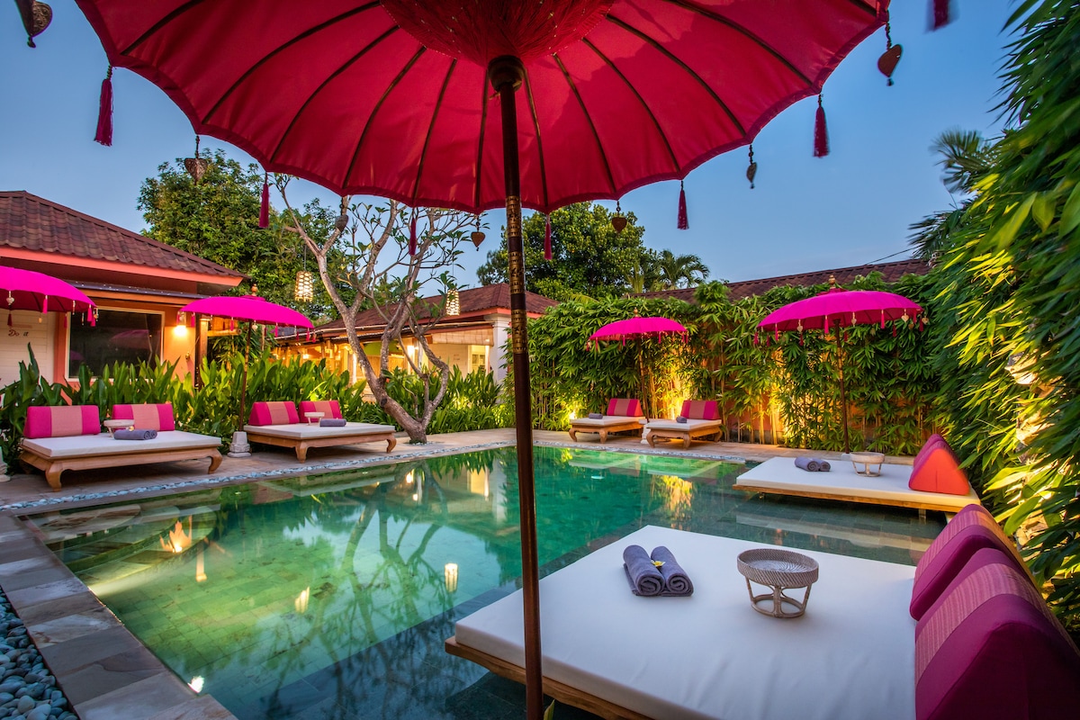 A vibrant pool area features oversized pink umbrellas providing shade over cushioned lounge chairs. Lush greenery encircles the space, contributing to a serene atmosphere. Soft lighting accentuates the pool, creating a calming evening ambiance for relaxation.
