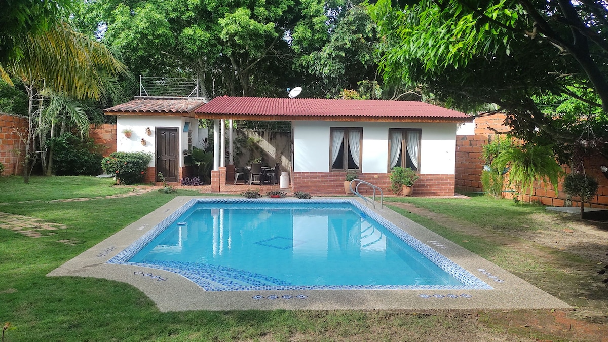 A tranquil outdoor area features a swimming pool, surrounded by neatly trimmed grass and landscaped greenery. The inviting water reflects the sunlight, while a nearby wooden structure with large windows provides a shaded space. Lush trees and flowering shrubs enhance the natural ambiance.