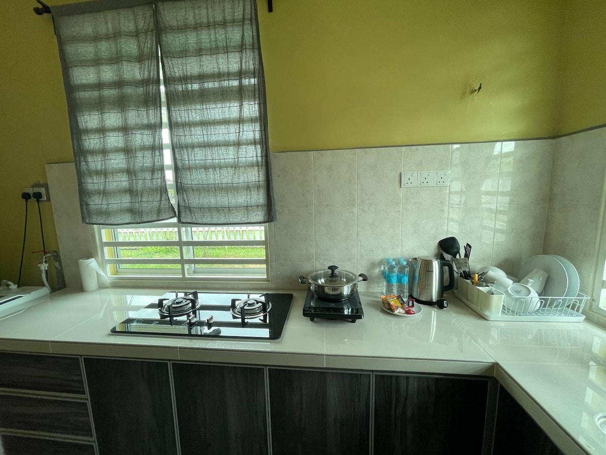 A functional kitchen area features a clean, modern countertop with a gas stove and cooking pot. Various kitchen utensils are neatly arranged, and a kettle is positioned beside bottled water. Natural light filters through a covered window, reflecting off the light-colored walls.