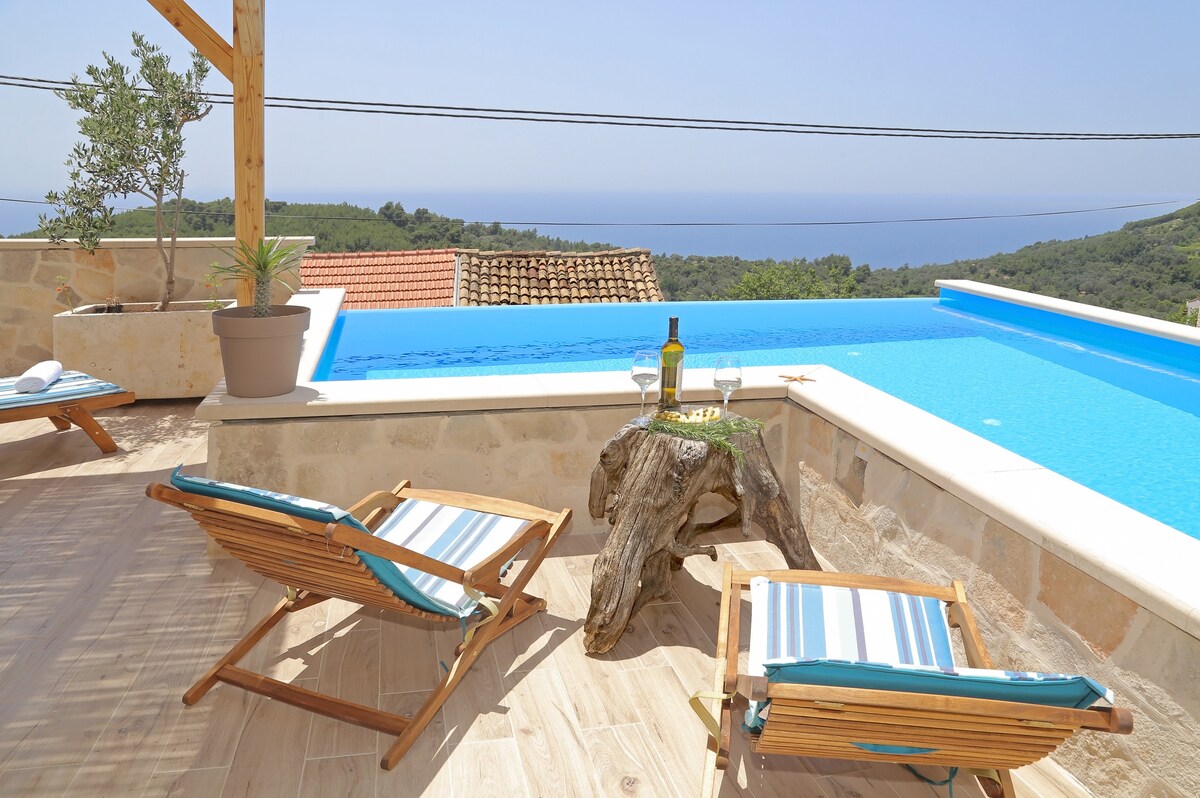 An inviting outdoor space includes two wooden lounge chairs positioned by the edge of a swimming pool. A small table made from natural wood holds a bottle of wine and snacks, overlooking scenic views of the Adriatic Sea and surrounding greenery.