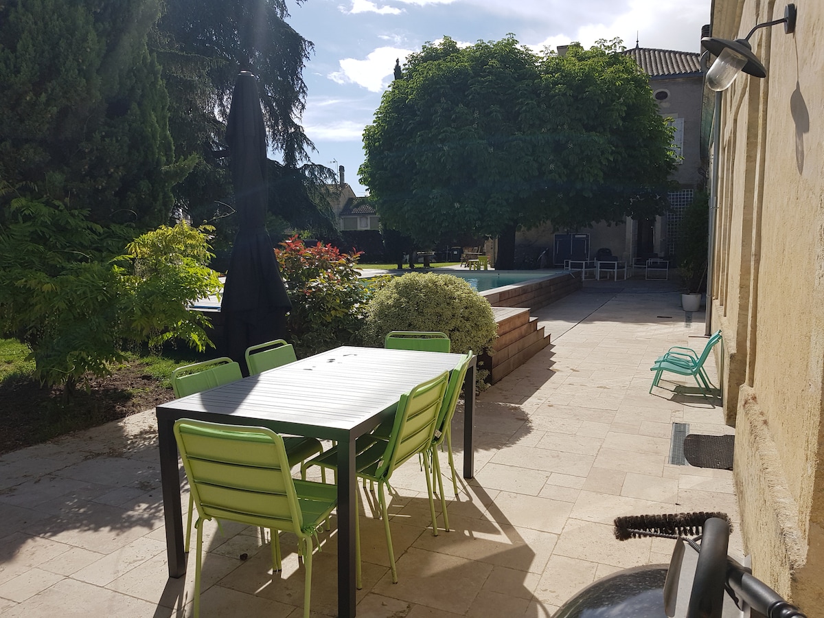 An outdoor area features a spacious dining table surrounded by green chairs, with a large tree providing shade nearby. A swimming pool is visible in the background, adding to the serene setting. Sunlight casts gentle shadows across the paving stones.