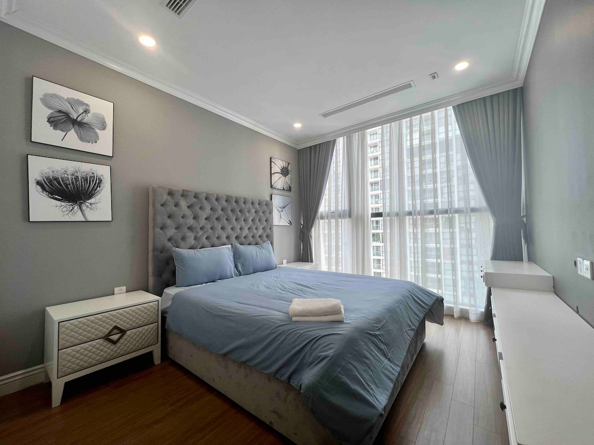 A spacious bedroom features a large upholstered bed with a light blue comforter. Two framed botanical prints adorn the wall above a bedside table. Natural light filters through floor-to-ceiling sheer curtains, creating a bright atmosphere. A minimalist dresser is positioned against the opposite wall.