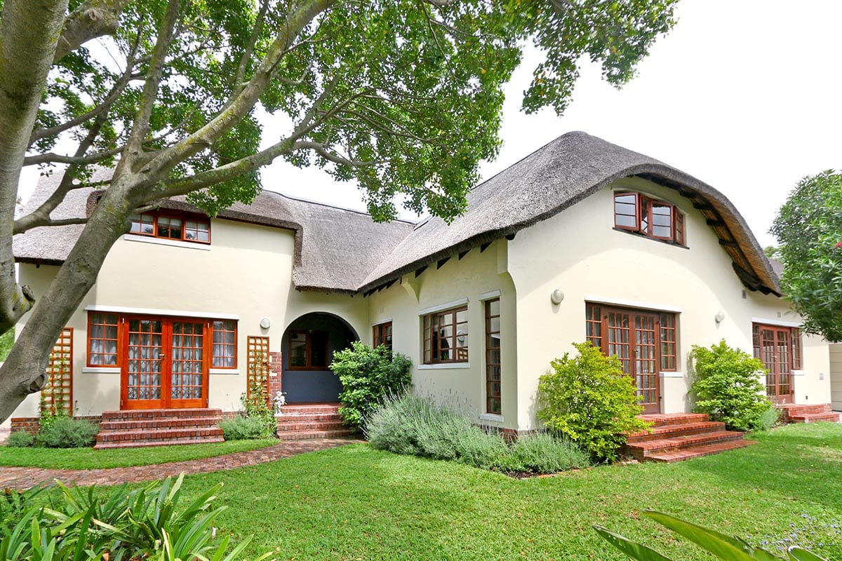 A thatched roof home is surrounded by a vibrant garden, featuring lush greenery and well-maintained shrubs. Large windows are framed in warm wood, enhancing the inviting façade. A pathway leads to the entrance, complemented by neatly arranged steps and natural stone accents.