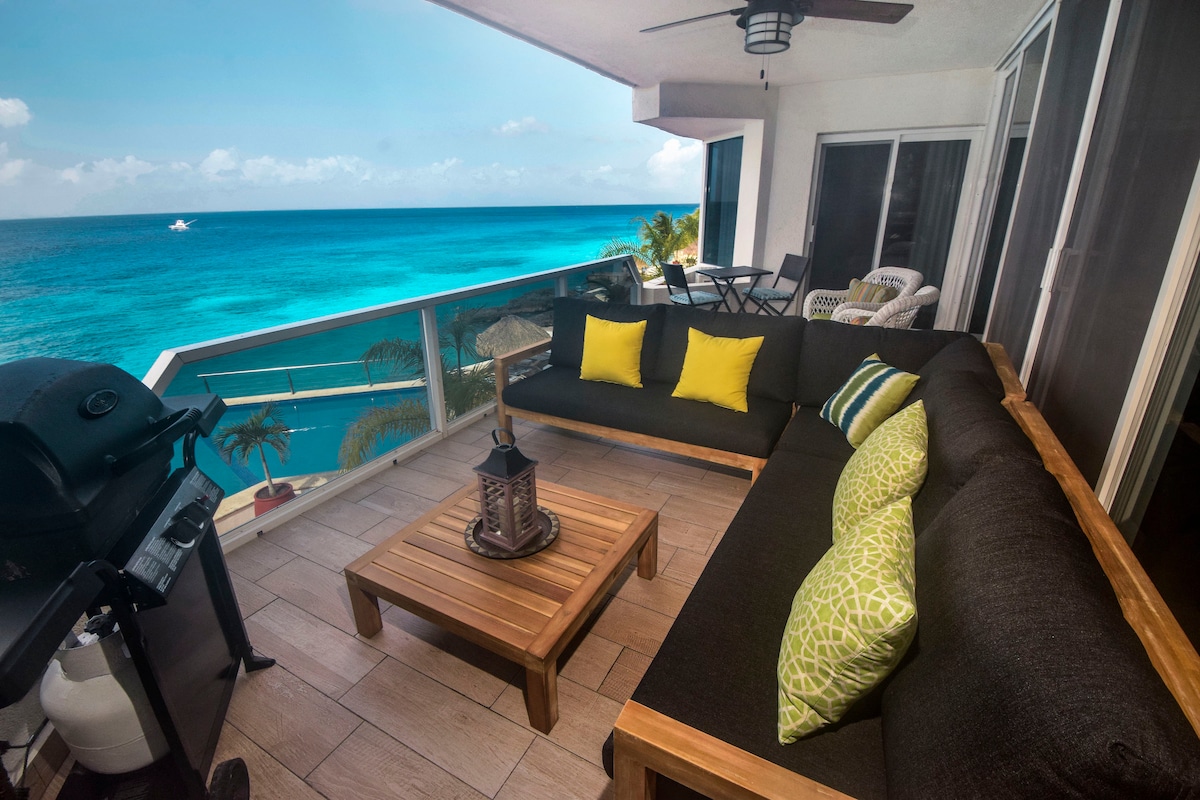 A spacious covered balcony is shown, featuring comfortable seating with bright yellow cushions and green accent pillows. A wooden table is placed in the center, complemented by a lantern. A grill is positioned to one side, offering ocean views over the turquoise water.