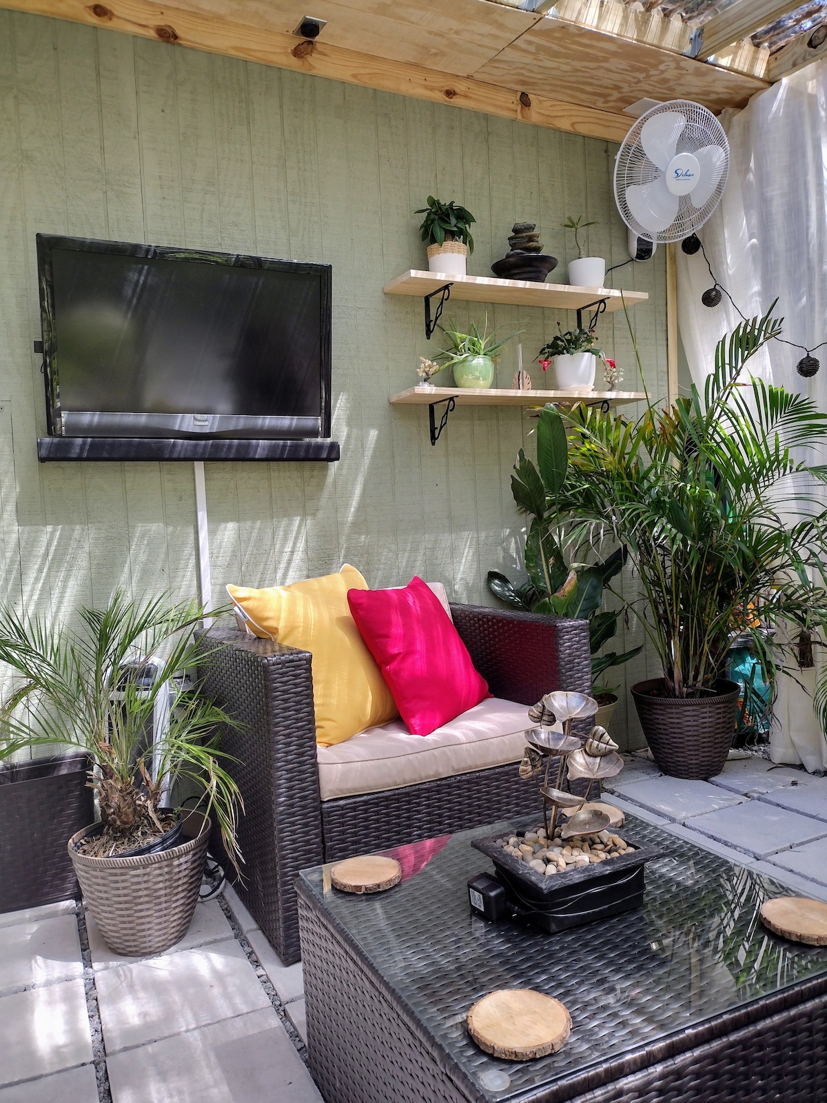 A cozy lounge area is surrounded by greenery. A comfortable seating arrangement is depicted, featuring a light-colored couch adorned with vibrant cushions. A glass coffee table with decorative coasters sits in front. A mounted TV and open shelving are visible against a green wall.