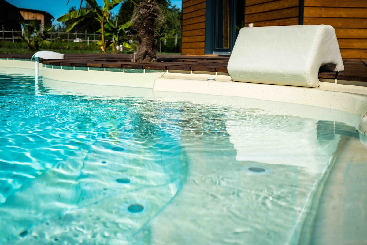 A close-up view of a serene swimming pool, showcasing clear blue water with gentle ripples. The pool's edge is lined with a soft white cushion for comfort. Surrounding foliage and a wooden deck offer a natural ambiance.