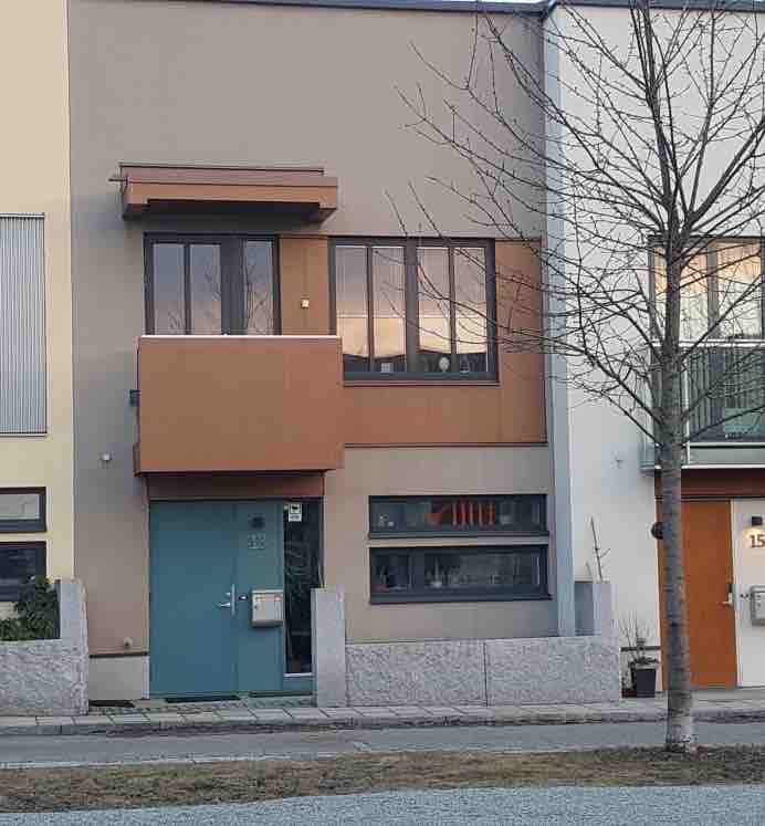 The townhouse features a contemporary façade with a combination of earth tones and large windows that provide natural light. The main entrance is marked by a turquoise door, surrounded by clean lines and a minimalist design, complemented by a small tree and gravel landscaping.