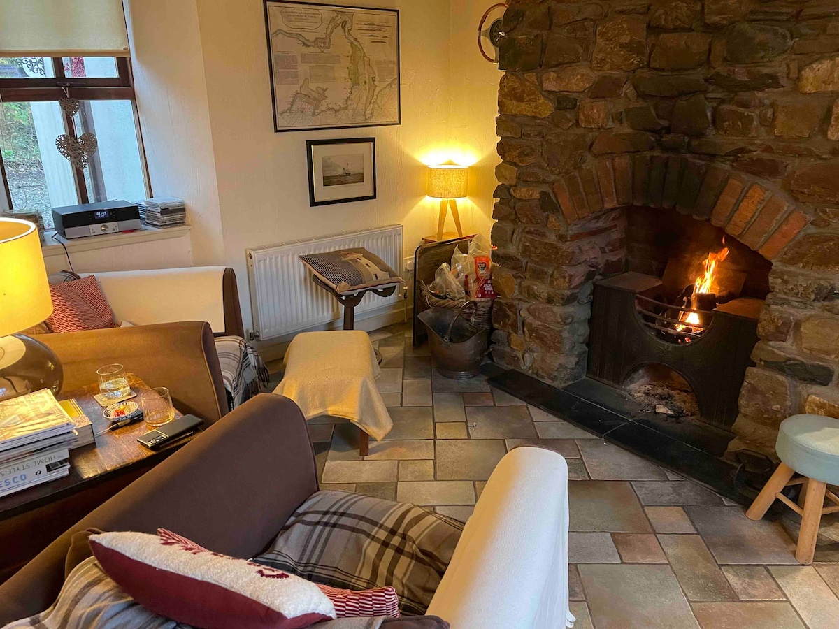A cozy sitting room features a stone fireplace with a warm fire. Light from a nearby lamp softly illuminates the space, highlighting comfortable seating and a rustic coffee table. Natural stone tiles add texture to the floor, complemented by an inviting atmosphere.