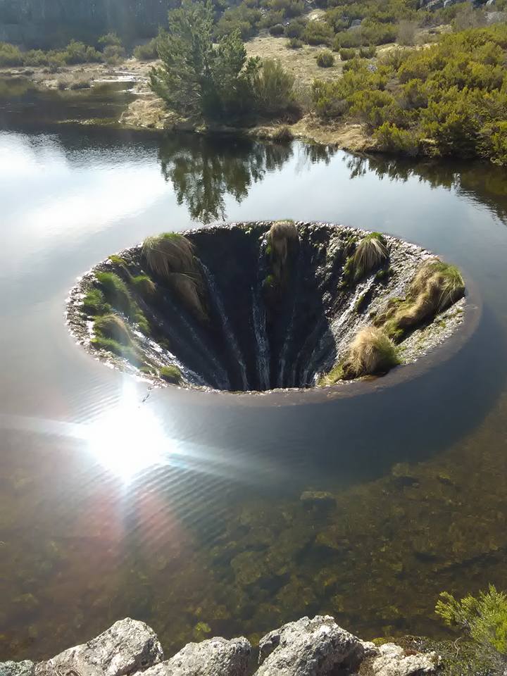 A unique natural formation is depicted, resembling a circular sinkhole surrounded by greenery. Water cascades over the edge, creating a small waterfall into the still body of water below. Sunlight reflects off the water's surface, adding a gentle sparkle.