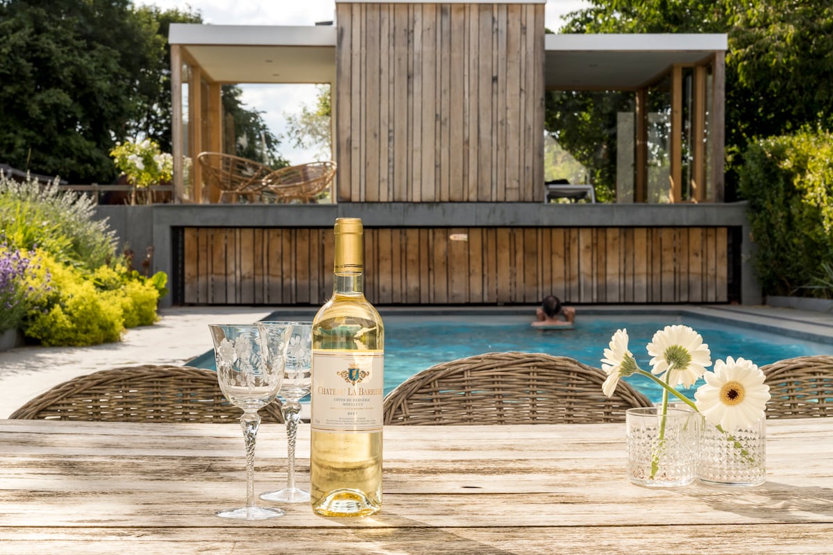 A wooden table is set with a bottle of white wine and two glasses, alongside a vase of flowers. In the background, a swimming pool is visible, with a person swimming. A wooden structure and lush greenery frame the scene, enhancing the tranquil atmosphere.