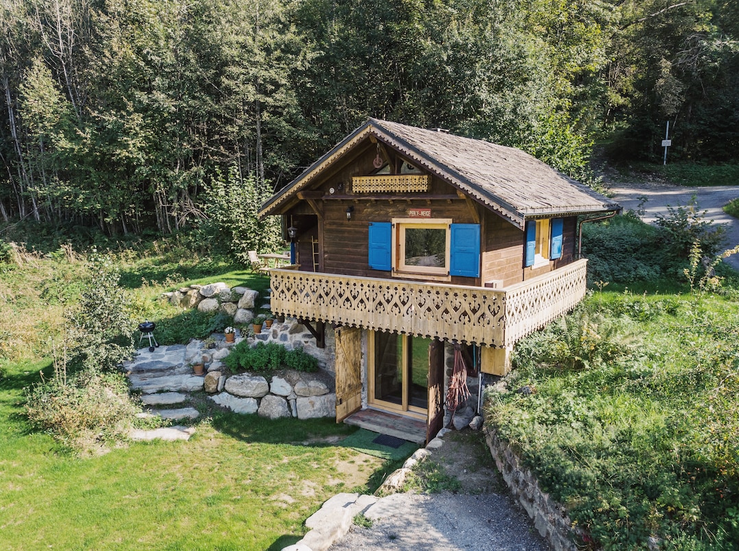 A charming mountain cabin is nestled among trees, featuring a wooden exterior with decorative balconies. Bright blue shutters contrast against the brown wood, while a well-kept garden surrounds the cabin. Nearby stone pathways lead to a welcoming entrance and outdoor barbeque area.
