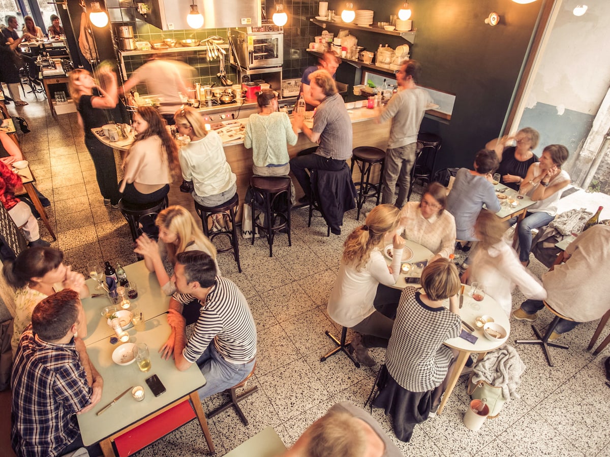 A bustling restaurant scene is depicted, featuring various tables occupied by guests enjoying meals. The bar area is visible in the background, with staff preparing food. Warm overhead lighting adds to the inviting atmosphere, while a blend of textures and colors is present throughout the space.