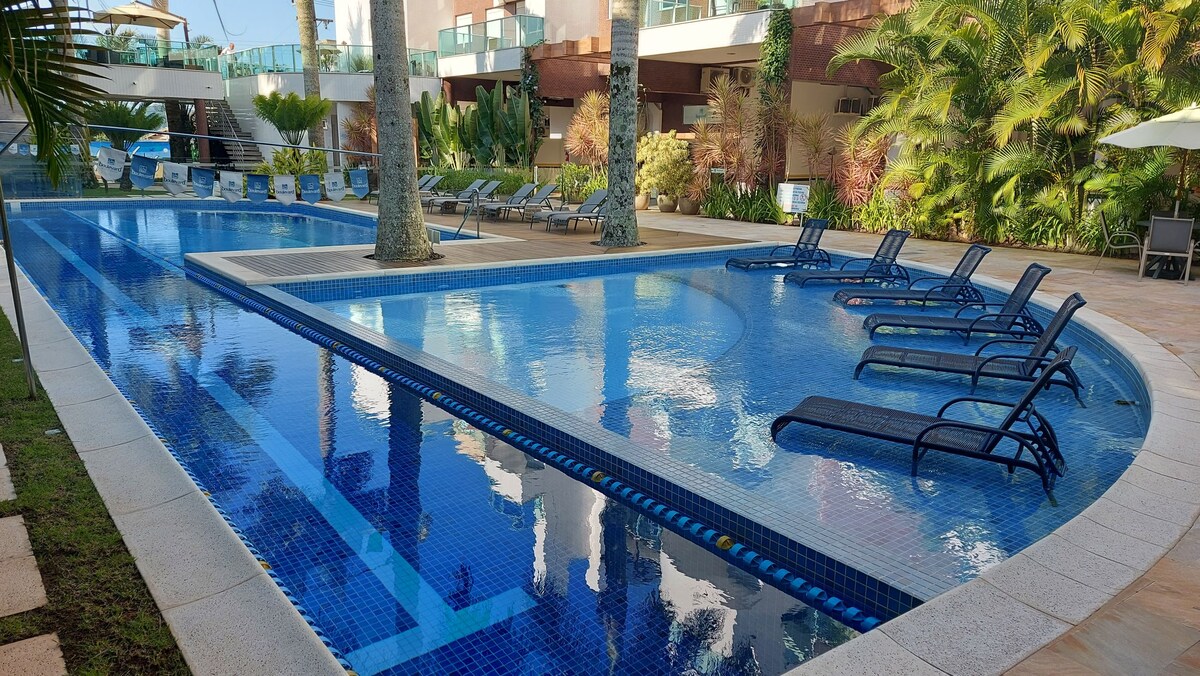 An inviting outdoor pool area is surrounded by lush greenery, featuring multiple lounge chairs positioned along the water's edge. The clear blue water reflects the surrounding palm trees and architecture, providing a serene space for relaxation.