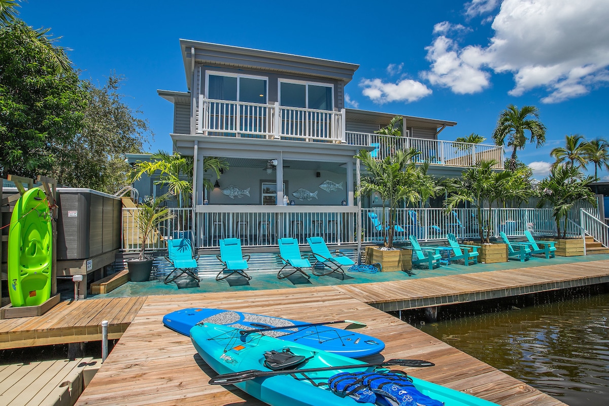 A waterfront view showcases a vibrant two-story building surrounded by greenery. Bright blue lounge chairs are positioned along the deck, with kayaks and paddle boards resting nearby. The expansive outdoor space, framed by tropical plants, invites relaxation and outdoor activities.