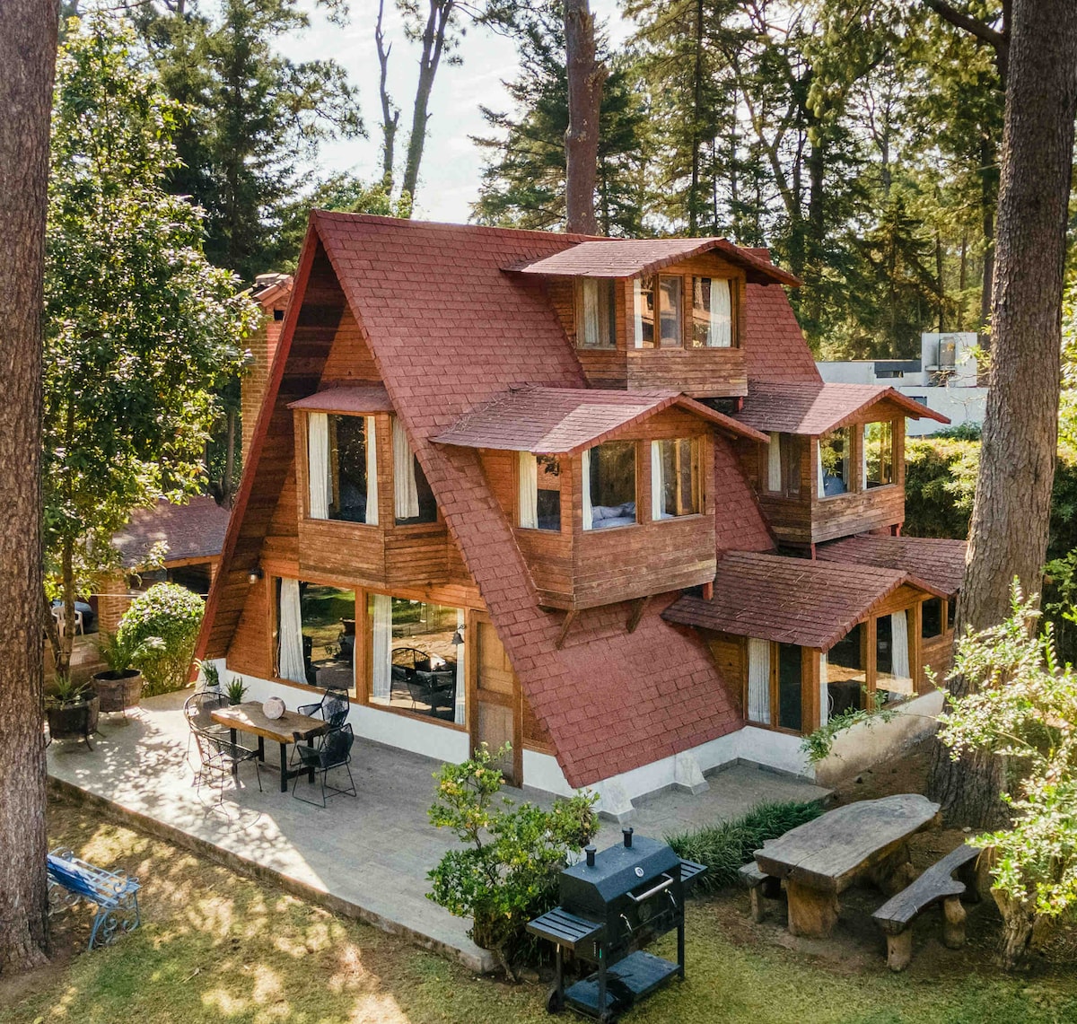 A multi-level cabin features a distinctive A-frame architecture, surrounded by tall trees. Large windows invite natural light, and outdoor seating accompanies a grill area on a tiled patio. The structure is nestled within a lush green landscape, enhancing the serene environment.
