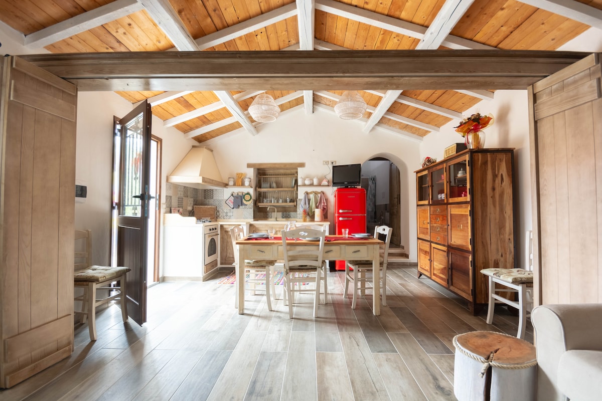 The open-concept living area features a wooden ceiling with beams, creating a spacious feel. A dining table is surrounded by white chairs, with a vibrant red refrigerator in the background. Natural light floods the space through a doorway and windows, enhancing the welcoming atmosphere.