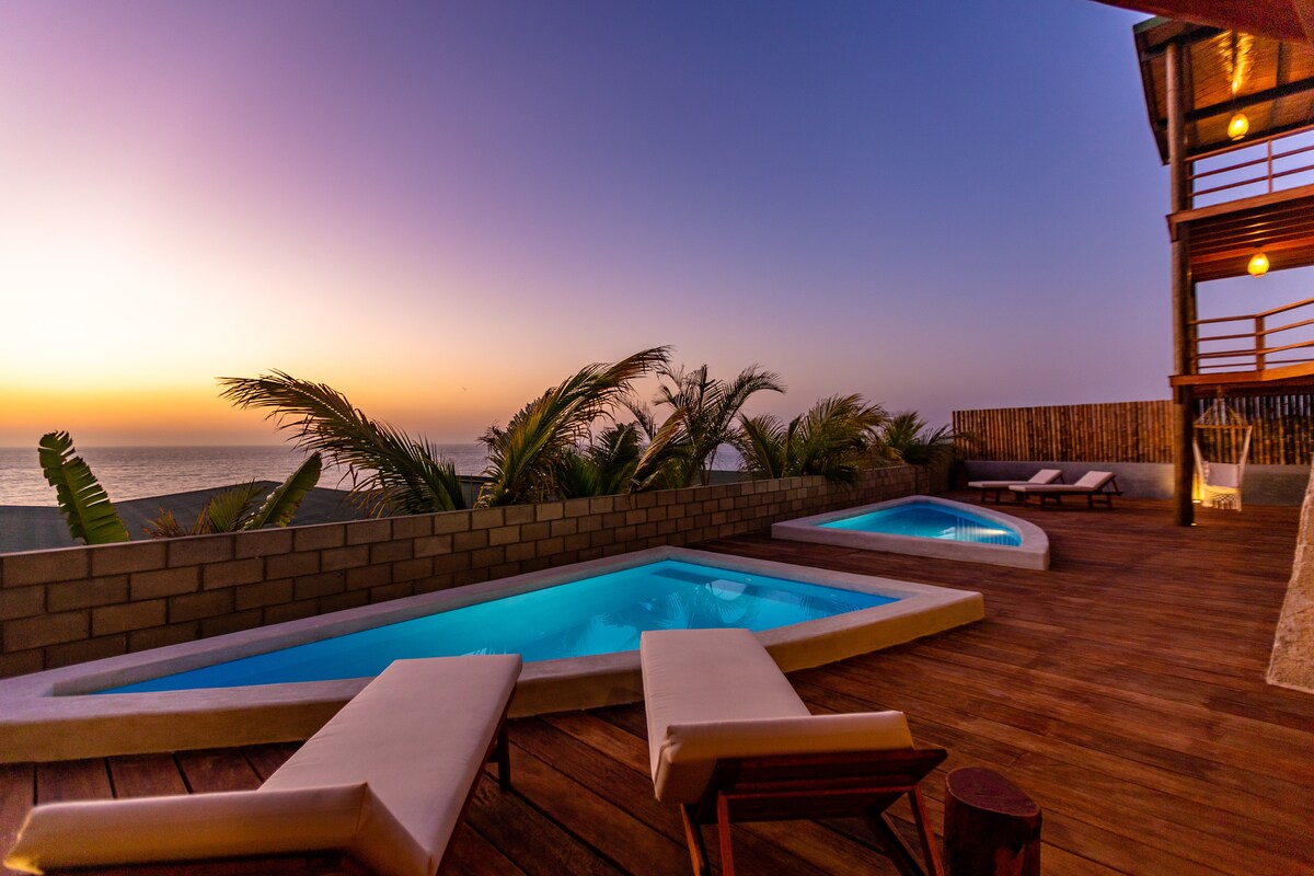 An outdoor deck features two inviting swimming pools under a twilight sky. Sun loungers are situated nearby, surrounded by tropical plants. The sea is visible in the background, creating a serene atmosphere as the sun sets.
