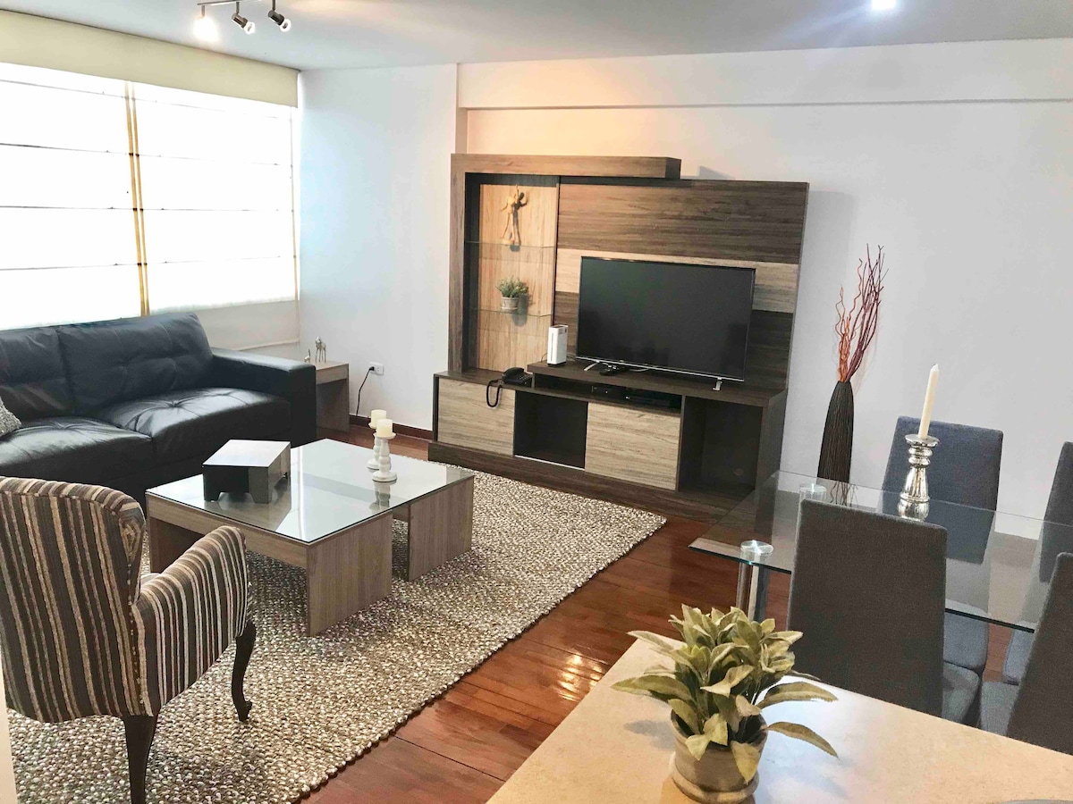 A spacious living area features a large black leather sofa and a modern entertainment center with a flat-screen TV. A rectangular glass coffee table sits on a textured area rug, surrounded by dark dining chairs. Natural light enters through large windows adorned with minimalistic coverings.