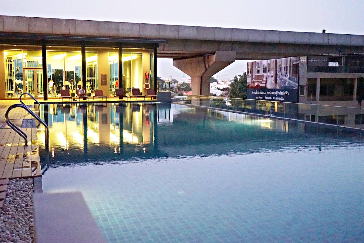 An expansive swimming pool reflects the evening sky, bordered by a wooden deck. The pool area is accompanied by a modern fitness center visible in the background, featuring large windows that showcase the interior. Soft lighting enhances the tranquil ambiance.