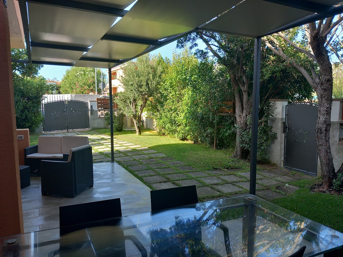 A covered outdoor seating area is visible, furnished with a glass dining table and black chairs. Beyond, a well-maintained garden features manicured grass and stone pathways leading towards a gate. Lush greenery and trees frame the space, creating a private and serene environment.