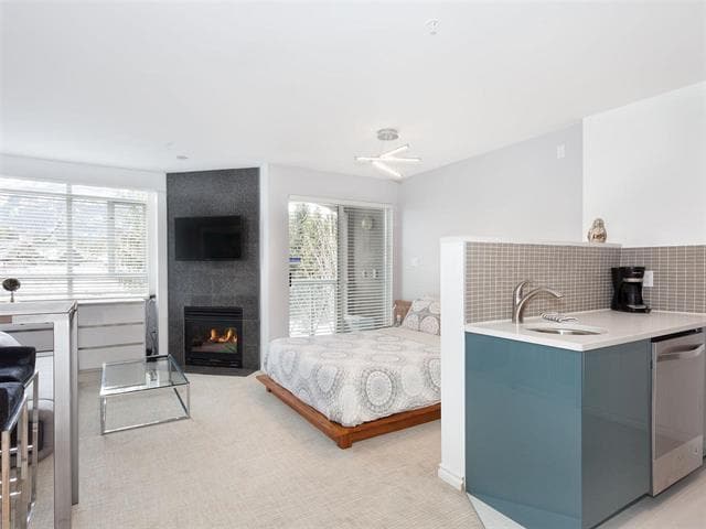 An open studio layout features a spacious bed near a cozy gas fireplace and a mounted TV. A modern kitchen area is visible with a microwave and refrigerator. Light enters through large windows, providing a bright and airy atmosphere.