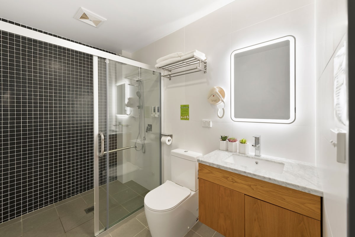 The bathroom features a glass-enclosed shower with modern fixtures and a contrasting black tiled wall. A sleek vanity with a marble countertop is complemented by a backlit mirror. Ample storage is provided by wooden cabinetry and shelves.