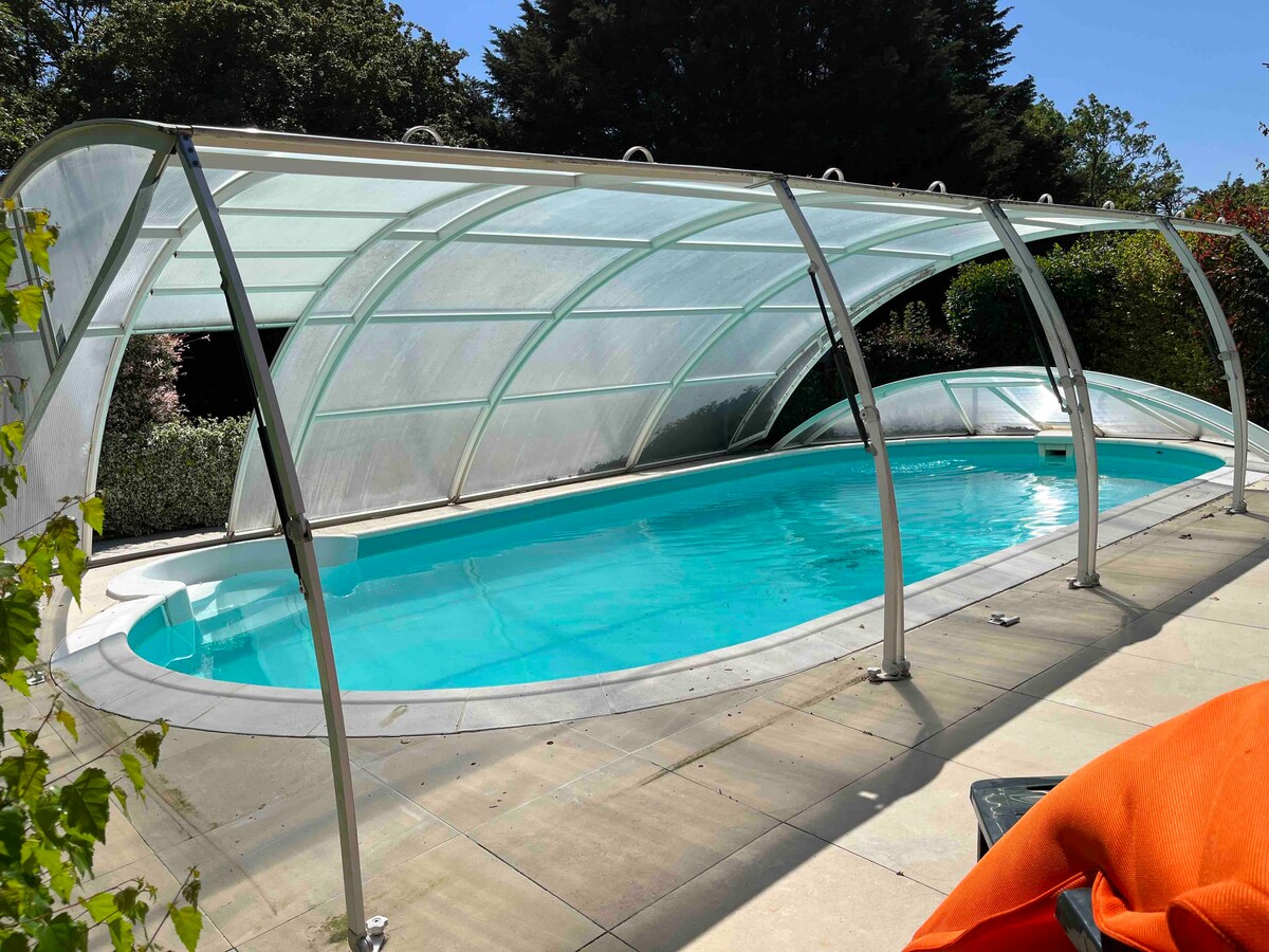 A covered swimming pool is visible, featuring a clear, rounded roof supported by a metal frame. The tranquil water reflects the surrounding greenery, with a spacious deck area made of stone tiles. Vibrant orange fabric is draped casually on the edge of the deck.