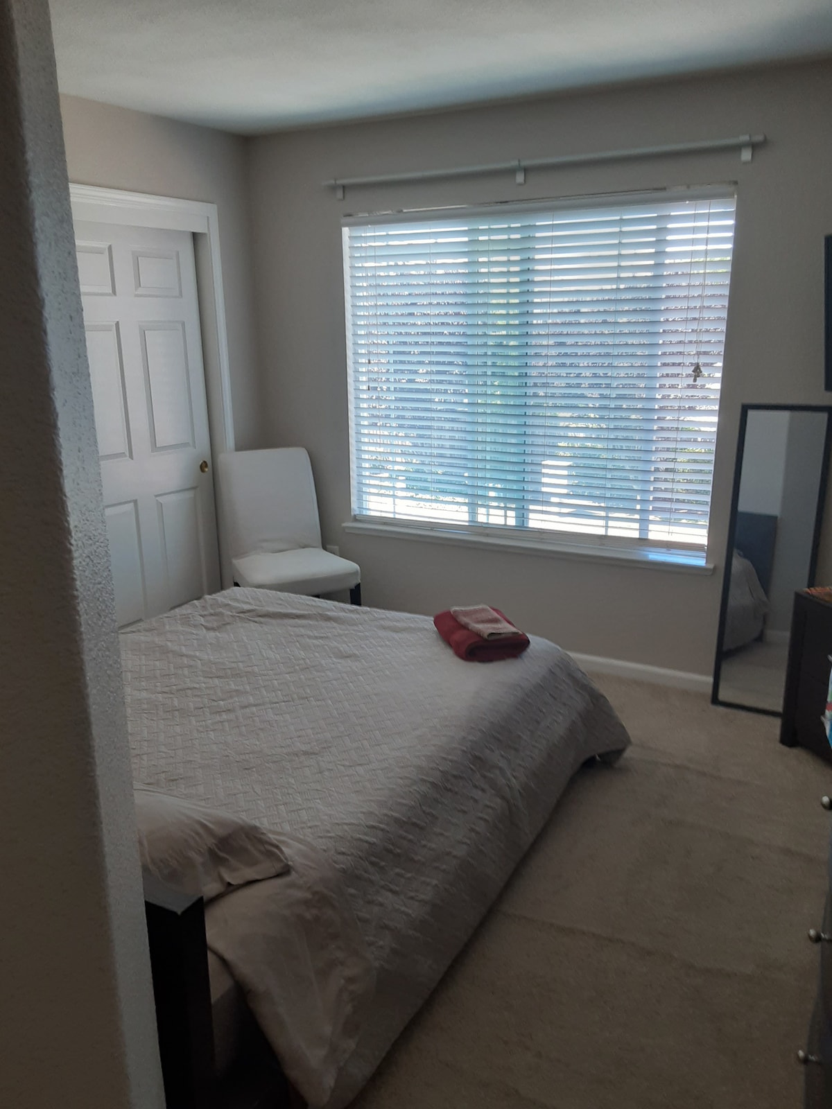 A bright room features a spacious bed with a light-colored coverlet, complemented by a neatly placed red towel. A chair sits beside the bed, and a mirror reflects the natural light from the window, which is dressed with white blinds.