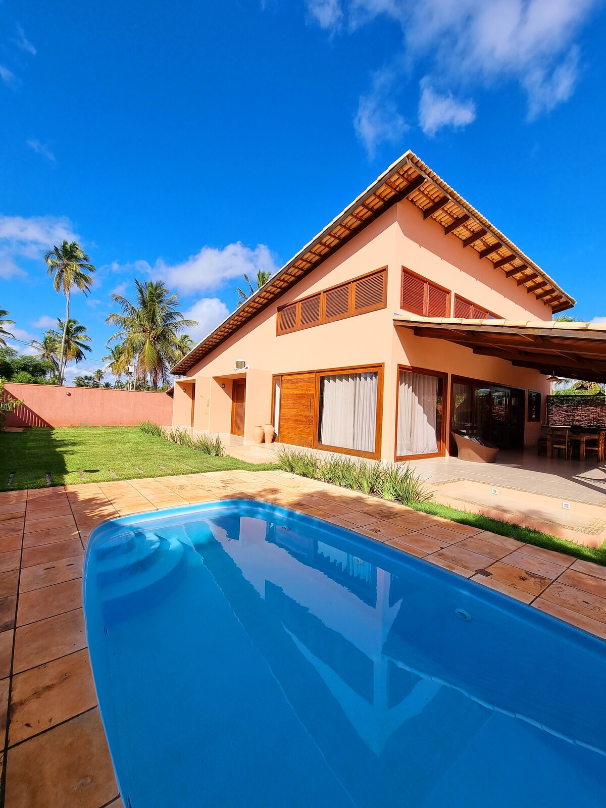 A modern two-story house is shown with a sloped roof and large windows. The image captures a small pool in the foreground, surrounded by a tiled terrace and lush greenery. Palm trees gently sway in the background under a clear blue sky.