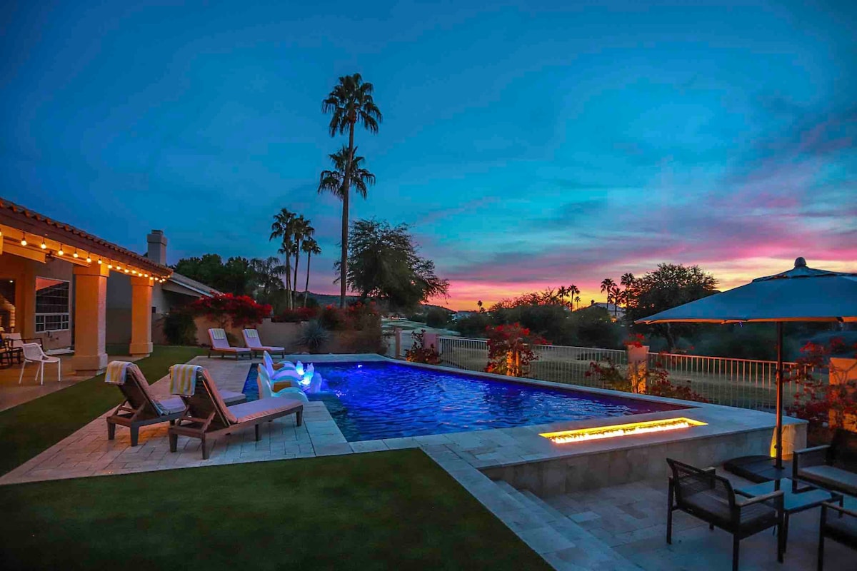 A serene pool area is highlighted by a heated pool with lounge chairs and a shallow baja shelf. Lush palm trees frame the space, while vibrant sunset colors paint the sky. An outdoor umbrella and seating area provide options for relaxation.