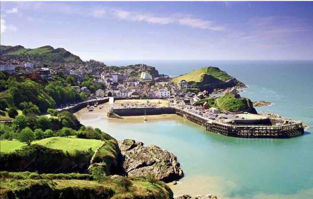 A panoramic view of Ilfracombe Harbour showcases its sandy beach and gently curving shoreline. Surrounding green hills rise in the background, while the blue waters of the coast meet the horizon. The quaint town's buildings nestle harmoniously against the landscape, reflecting a charming seaside atmosphere.