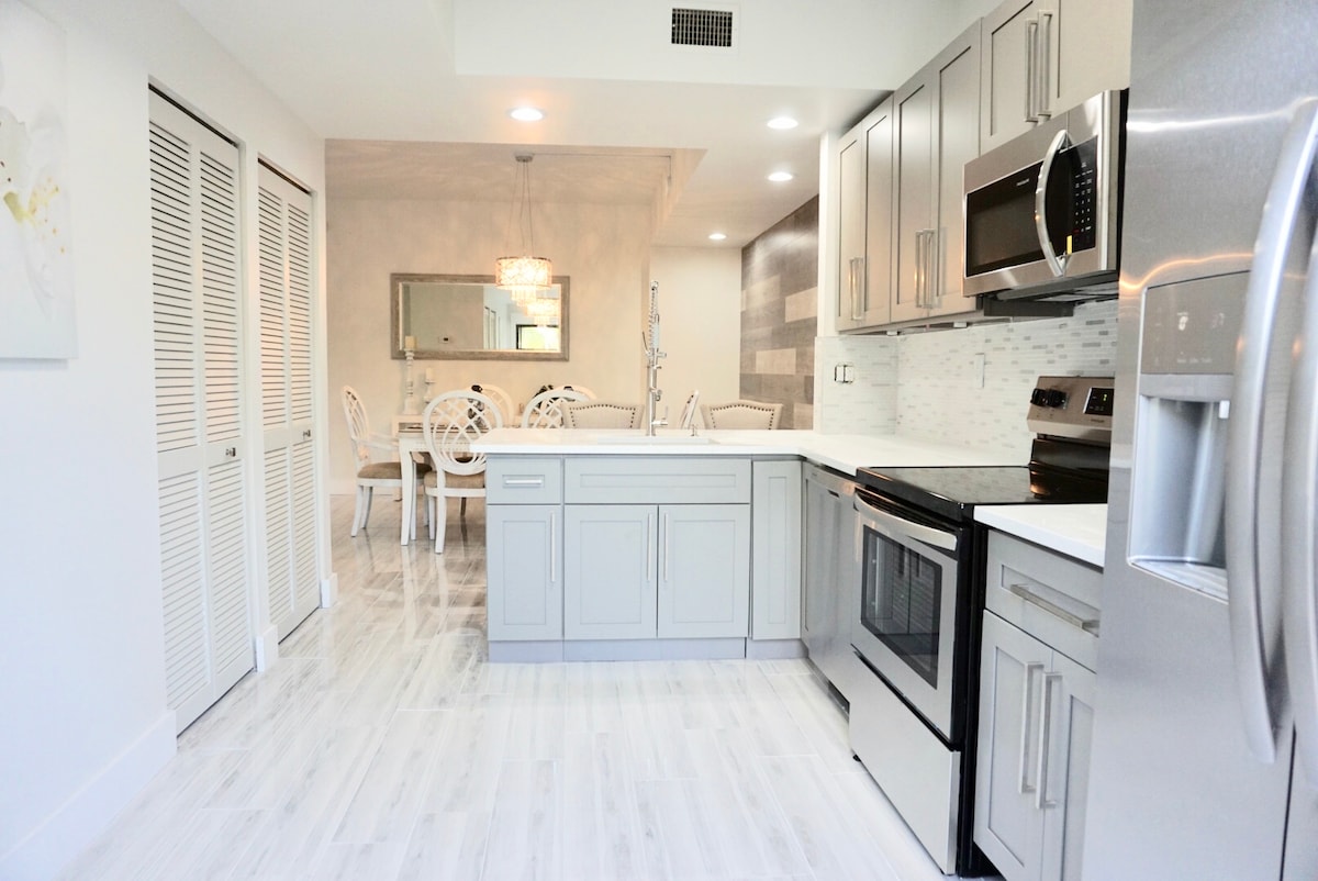 A modern kitchen is featured with gray cabinetry and stainless steel appliances. A spacious white countertop provides ample preparation space, while a dining area is visible in the background, showcasing a table with elegant white chairs. Soft lighting enhances the contemporary atmosphere.
