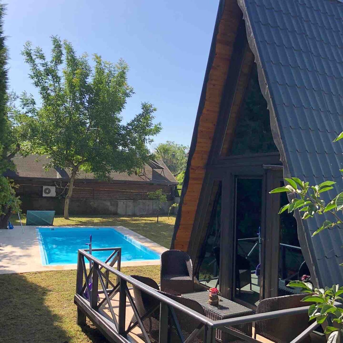 A charming A-frame structure is shown alongside a private pool, surrounded by lush greenery. The spacious deck features comfortable seating and a small table, providing a relaxing outdoor space. Sunlight highlights the pool's clear blue water, inviting guests to enjoy the tranquil atmosphere.