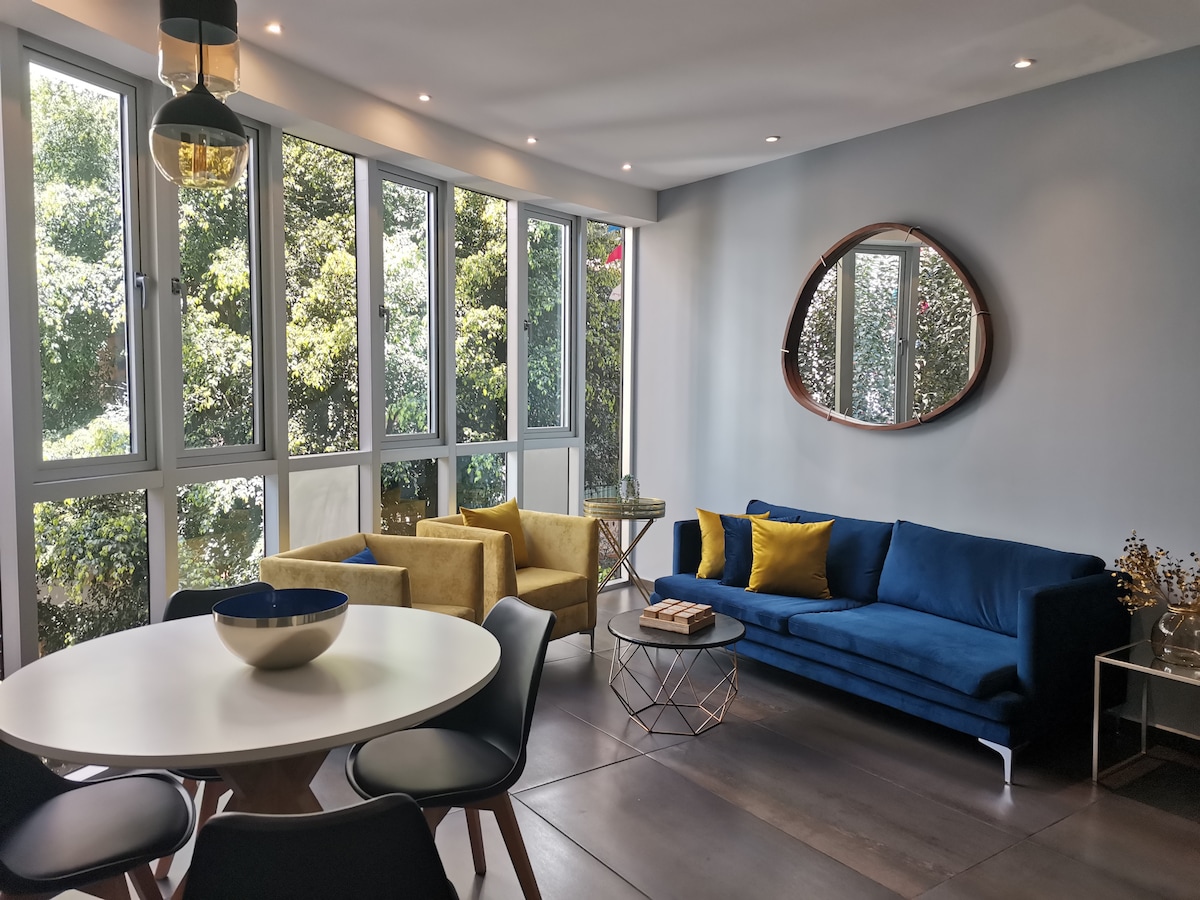 A living area features large windows allowing natural light to fill the space. The seating arrangement includes a blue sofa, yellow armchairs, and a round dining table. A decorative mirror is positioned on the wall, enhancing the open atmosphere of the room.