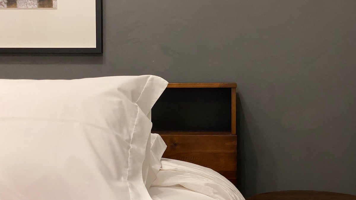 A close-up of a spacious bed features crisp, white bedding and plush pillows. A dark wooden headboard complements the neutral grey wall, while a nightstand adds a touch of warmth. Natural light softly illuminates the area.