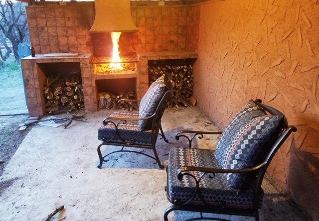 A covered patio area features two metal chairs with patterned cushions, positioned beside a stone fireplace. The fireplace, designed for grilling, showcases stacks of firewood on either side. Soft lighting from the flames enhances the warm ambiance of the space.