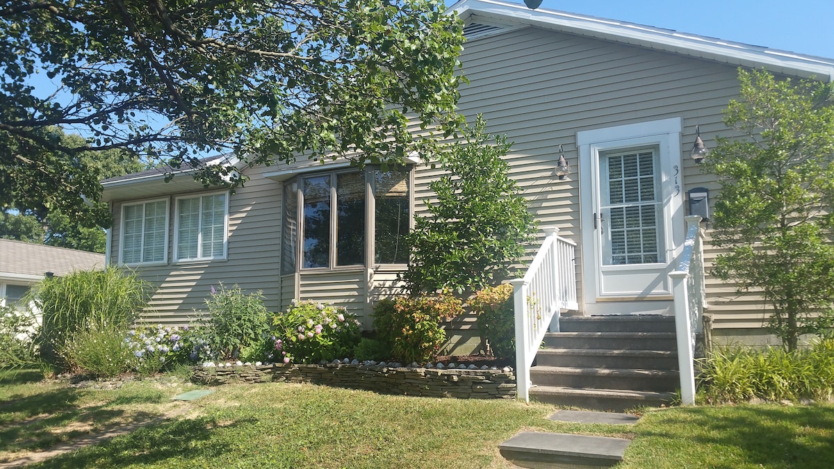 The exterior of the beach cottage features light gray siding with white trim and multiple windows. A set of steps leads to a welcoming entrance, bordered by landscaping that includes bushes and flowering plants. Sunlight brightens the scene, showcasing the cottage's inviting facade.