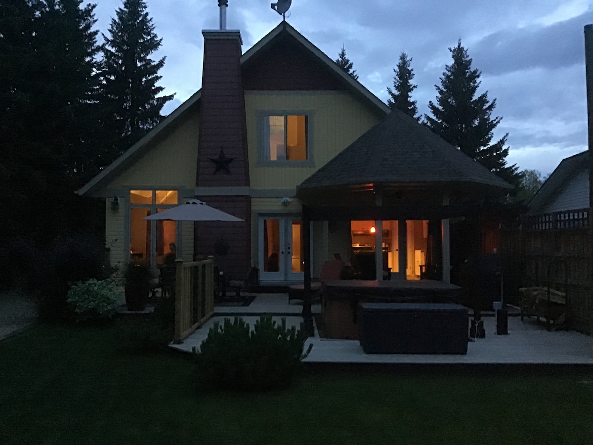 A charming lake home is set against a twilight backdrop, featuring warm light glowing from the windows. A covered outdoor area is visible, with a hot tub and seating arrangements on a wooden deck, surrounded by a lush lawn and tall trees.
