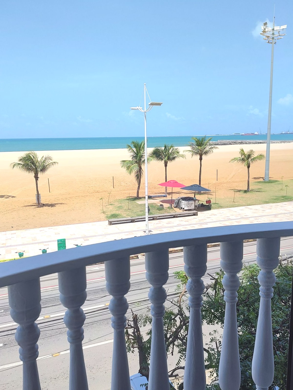 A view from the balcony overlooks a sandy beach with gentle waves in a vibrant blue ocean. Palm trees sway lightly in the breeze, and shaded areas provide places to relax. A smooth road runs beside the beach, connecting to nearby amenities.