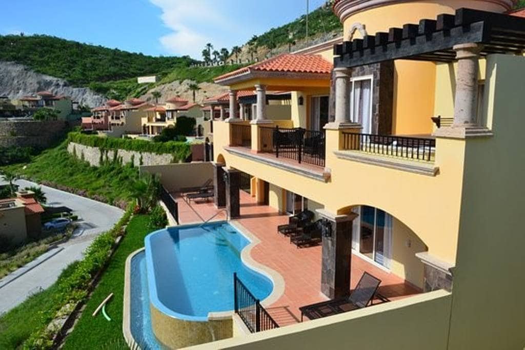 An elegant villa is situated on a hillside, featuring a private pool with a curved edge and surrounded by a spacious patio. The covered balcony provides shaded seating, and the landscape showcases beautiful greenery and distant terraces of neighboring villas.