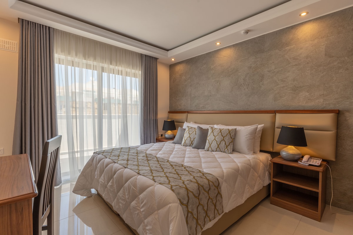 A double room is presented with a spacious bed neatly covered in a white quilt. Two bedside lamps flank the bed on wooden nightstands. Floor-to-ceiling curtains allow natural light to filter in, complementing the neutral tones of the decor.