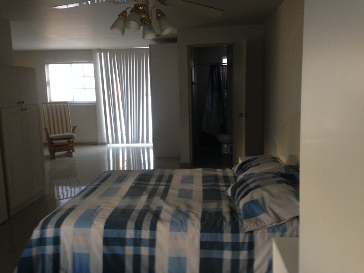 A spacious sleeping area features a comfortably made bed with a blue and white plaid bedding set. Natural light enters through a large window with vertical blinds. A rocking chair is positioned in one corner, while an adjoining room contains a shower and a toilet behind a door.