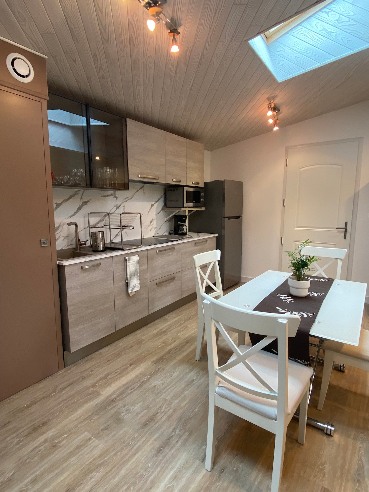 A modern kitchen area features light wood cabinetry and a marble backsplash. A dining table with white chairs sits in front of a large window, allowing natural light to fill the room. Appliances, including a refrigerator and a microwave, are visible.