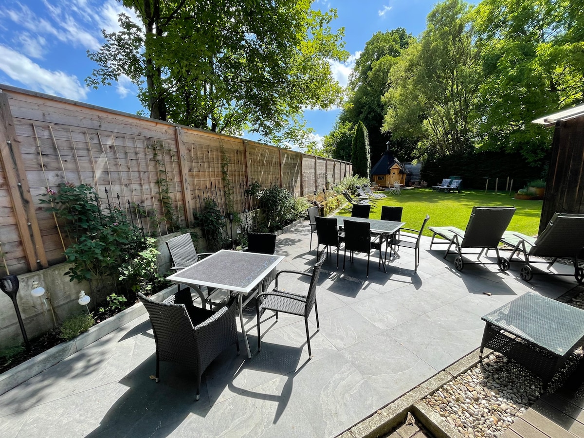 An outdoor terrace presents a spacious layout featuring several black wicker tables and chairs. Sun loungers are arranged around the area, surrounded by greenery and illuminated by natural light, creating an inviting space for relaxation.