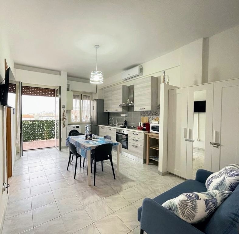 An open kitchen and dining area is shown, featuring a dining table set for four. Modern appliances and cabinetry are visible, along with a comfortable blue sofa. Natural light enters through a nearby balcony door, complemented by greenery from outside.