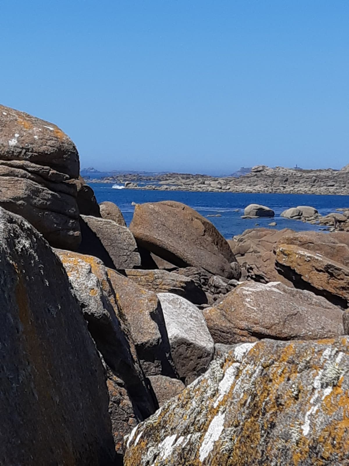 A rocky shoreline is depicted, with large natural stones scattered throughout the foreground. The smooth surfaces of the rocks are interspersed with patches of lichen. In the background, a calm blue sea stretches towards the horizon under a clear sky.