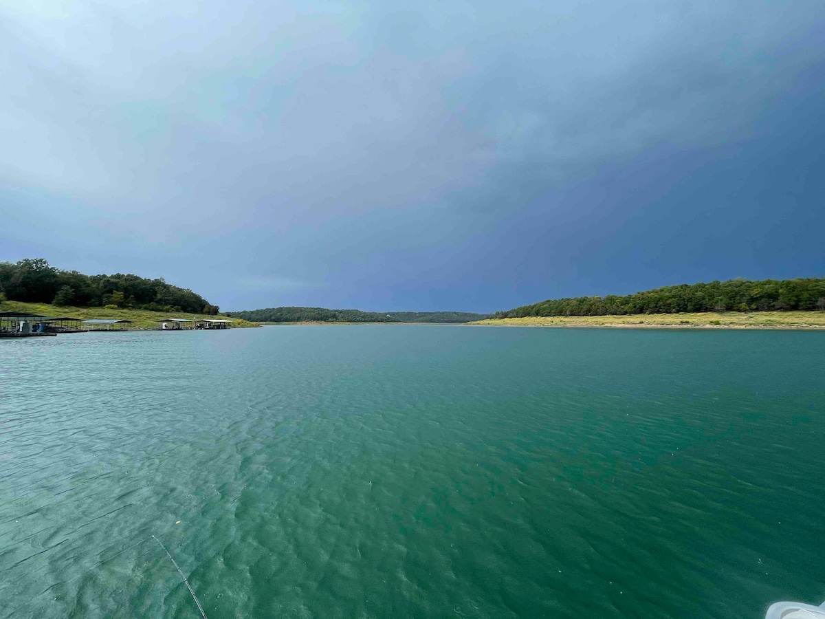 A calm water body stretches across the image, reflecting a serene blue-green hue. Lush green hills line the shore, under an expansive sky with varying shades of gray. Distant docks are visible along the edge, contributing to the tranquil lakeside landscape.