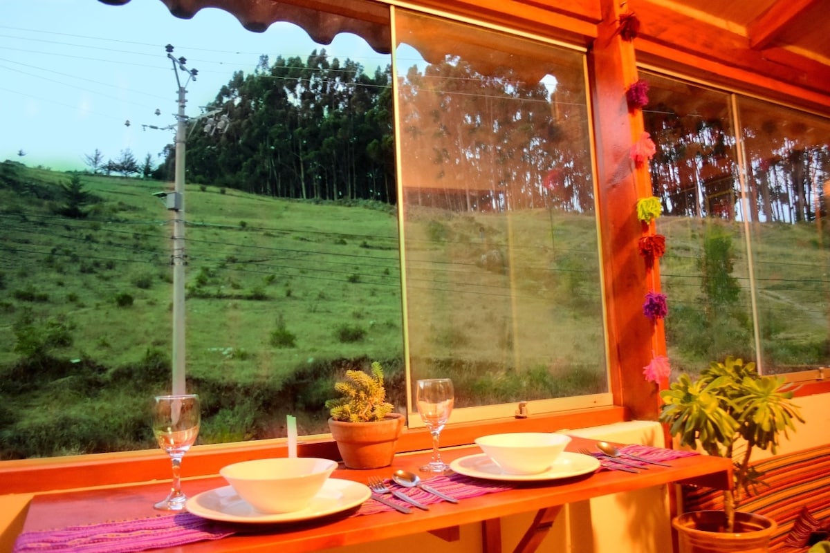 A dining area is presented with a set table featuring white plates and glasses. Large windows allow views of the lush green hillside and tall eucalyptus trees, creating a serene backdrop. A potted plant is visible on the table, adding a touch of nature.