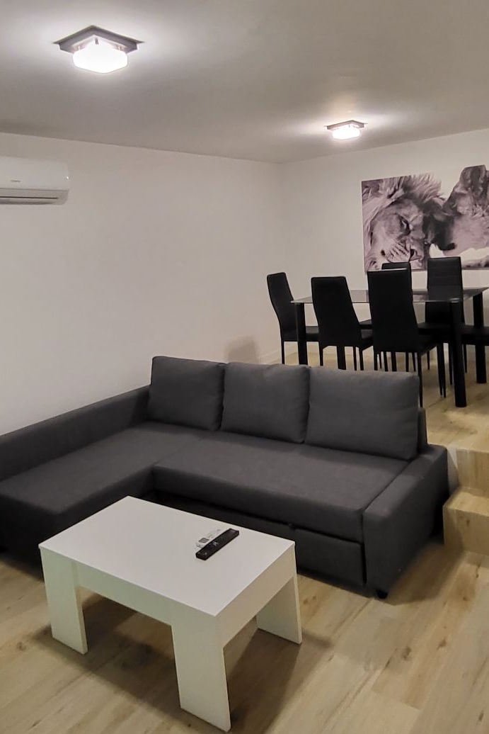 A modern living area features a grey sectional sofa paired with a minimalist white coffee table. In the background, a dining area showcases a glass-top table surrounded by six black chairs, complemented by a black and white artwork on the wall.