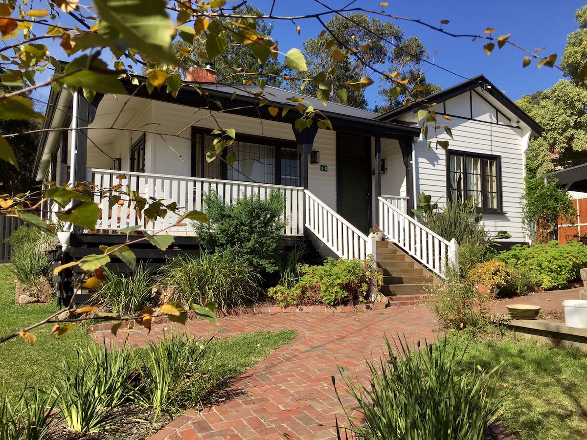 A charming country cottage is surrounded by lush greenery, featuring a brick pathway leading to a welcoming entrance. The front porch is highlighted by white railings and steps, showcasing the traditional architecture and creating an inviting atmosphere for guests.